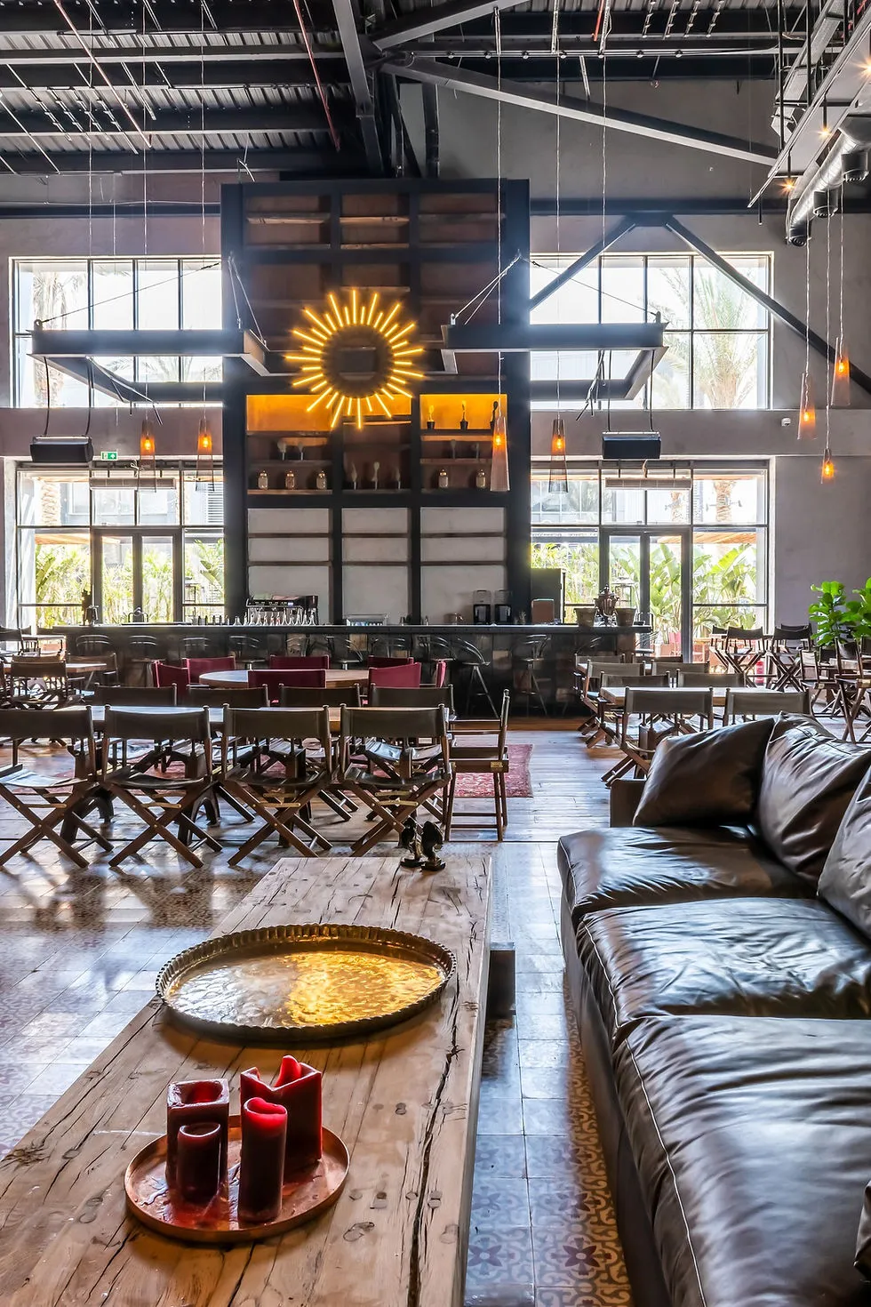 A spacious, modern lounge area with large windows, a black leather sofa, a wooden coffee table with red candles, and a bar in the background featuring a decorative sunburst neon sign.